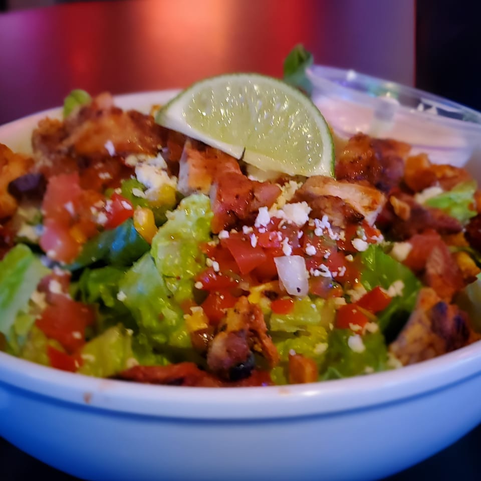 A close-up of a vibrant chicken salad with lettuce, tomatoes, corn, cheese, and a lime wedge.
