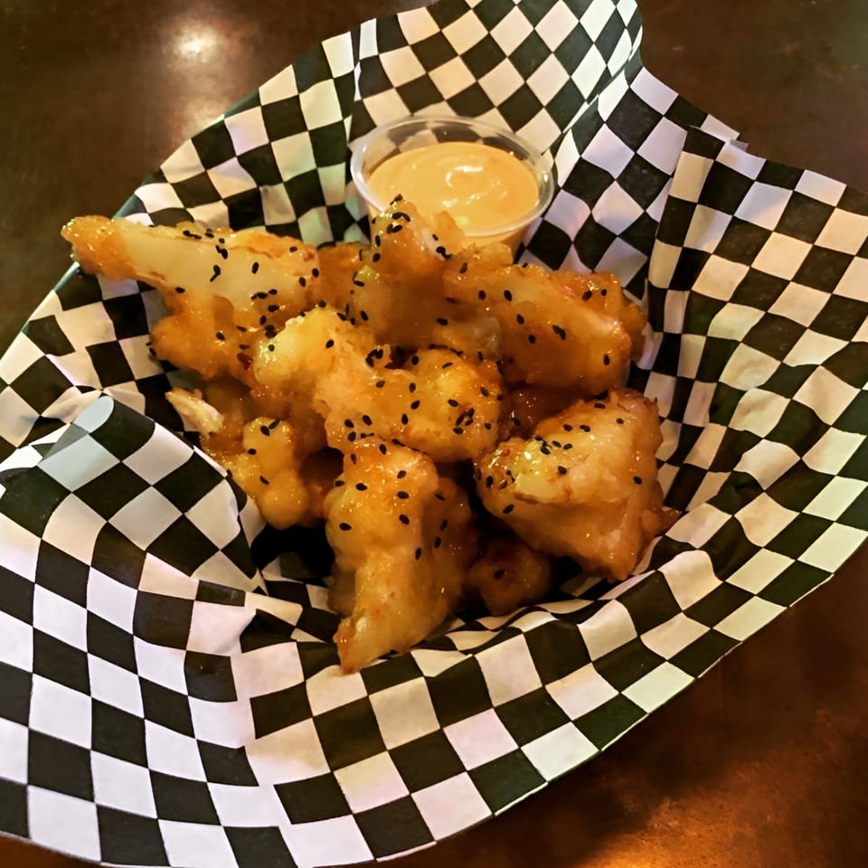 Crispy fried cauliflower bites with black sesame seeds and dipping sauce in a checkered paper basket.